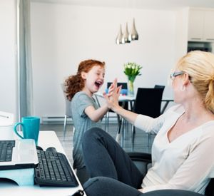 Eine Frau sitzt vor einem Computer. Neben ihr ist ein Kind, die beiden schauen sich an und klatschen sich ab. 
