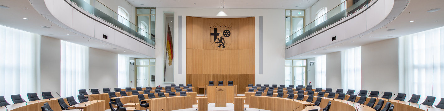 Plenarsaal des Landtags Rheinland-Pfalz. Stuhlreihen sind kreisförmig vor Tischreihen angeordnet. Im Hintergrund ist das rheinland-pfälzische Wappen neben einer großen Flagge. 