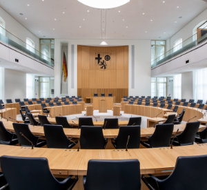 Plenarsaal des Landtags Rheinland-Pfalz. Stuhlreihen sind kreisförmig vor Tischreihen angeordnet. Im Hintergrund ist das rheinland-pfälzische Wappen neben einer großen Flagge. 