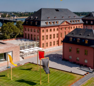Außenansicht des Landtags Rheinland-Pfalz. Ein großes Gebäude mit einem etwas kleineren vornedran. Im Hintergrund sind eine Brücke sowie ein Fluss.