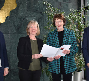 Eine Gruppe von vier Personen. Drei Frauen und ein Mann stehen vor einer Wand. Die beiden Frauen in der Mitte halten eine Mappe.