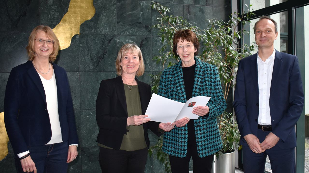 Eine Gruppe von vier Personen. Drei Frauen und ein Mann stehen vor einer Wand. Die beiden Frauen in der Mitte halten eine Mappe.