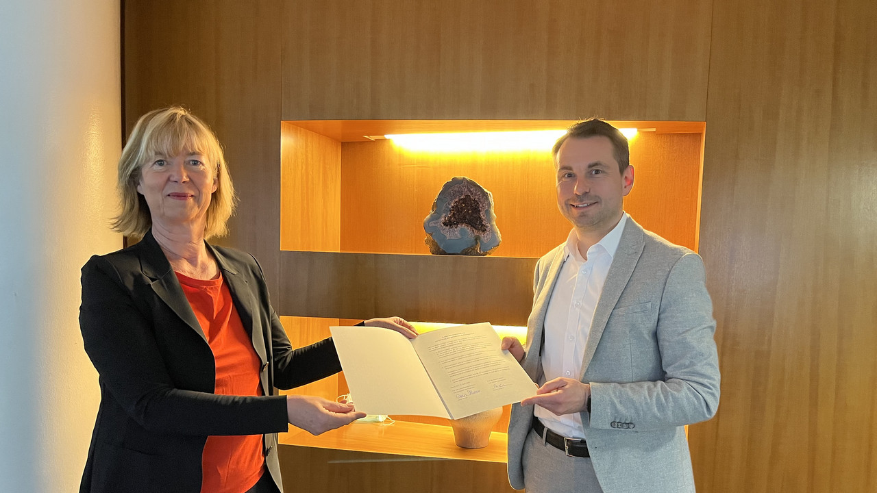 Zwei Personen stehen vor einem Regal und halten eine Vereinbarung in einer Mappe in der Hand. Sie schauen in die Kamera.