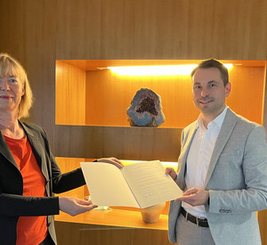 Zwei Personen stehen vor einem Regal und halten eine Vereinbarung in einer Mappe in der Hand. Sie schauen in die Kamera.