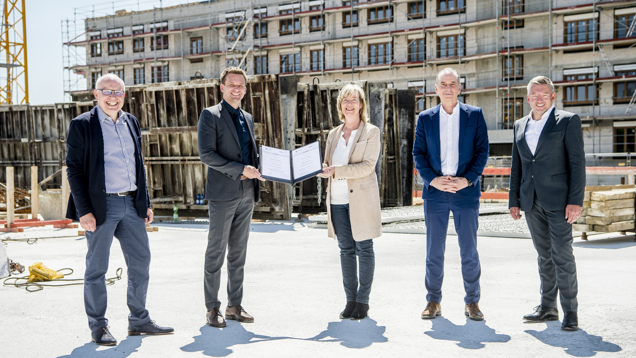 Fünf Menschen stehen vor einem Neubau, der sich noch im unverputzten Rohbau befindet. Die Frau in der Mitte überreicht dem Mann links von ihr einen Förderbescheid.