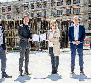 Fünf Menschen stehen vor einem Neubau, der sich noch im unverputzten Rohbau befindet. Die Frau in der Mitte überreicht dem Mann links von ihr einen Förderbescheid.