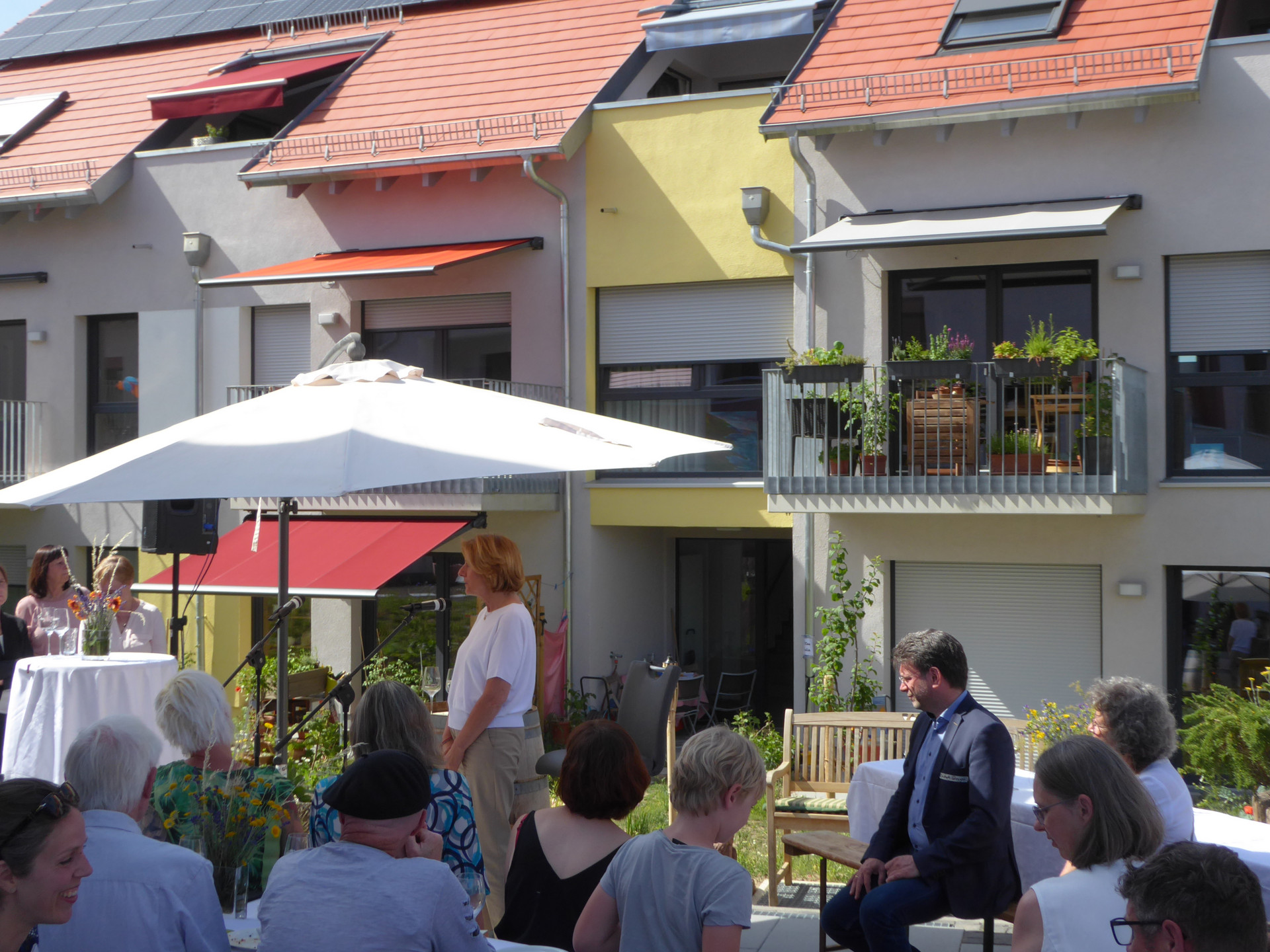 Ministerpräsidentin Malu Dreyer bei der Rede unter einem Sonnenschirm, davor sitzend Zuhörer vor dem Hintergrund eines Wohngebäude