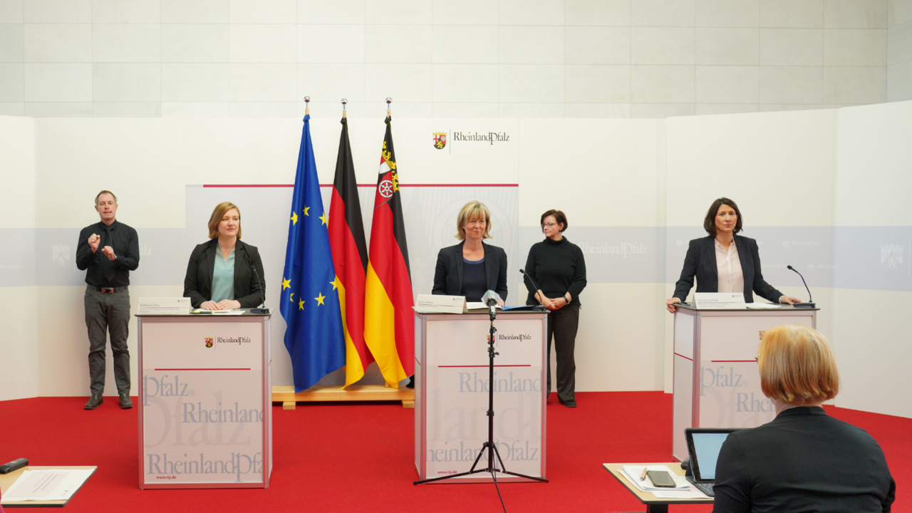 Drei Frauen stehen an Redepulten. Sie lächeln in die Kamera. Im Hintergrund sind zwei Gebärdendolmetschende platziert. 