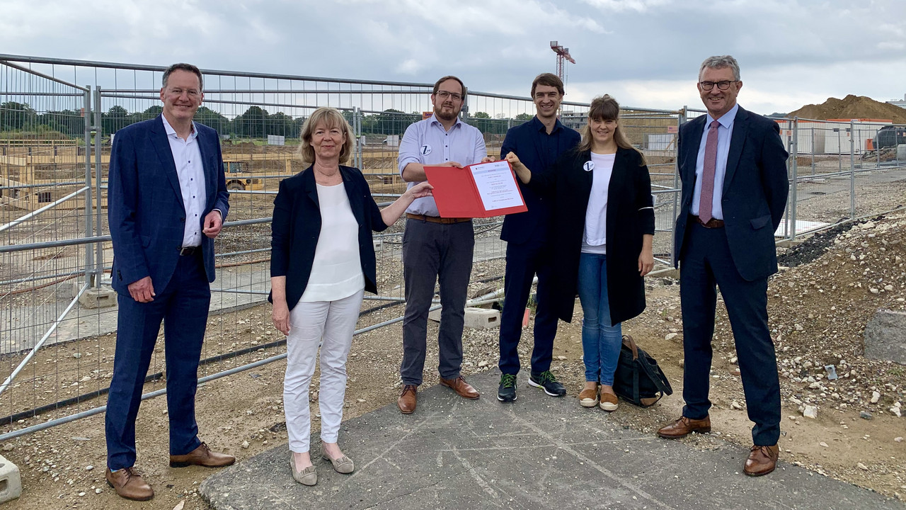 Eine Gruppe von sechs Menschen steht auf einer Baustelle. Davon sind zwei Frauen. Die beiden Frauen halten in der Mitte eine Mappe mit einem Förderbescheid in die Höhe.