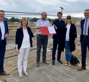 Eine Gruppe von sechs Menschen steht auf einer Baustelle. Davon sind zwei Frauen. Die beiden Frauen halten in der Mitte eine Mappe mit einem Förderbescheid in die Höhe.