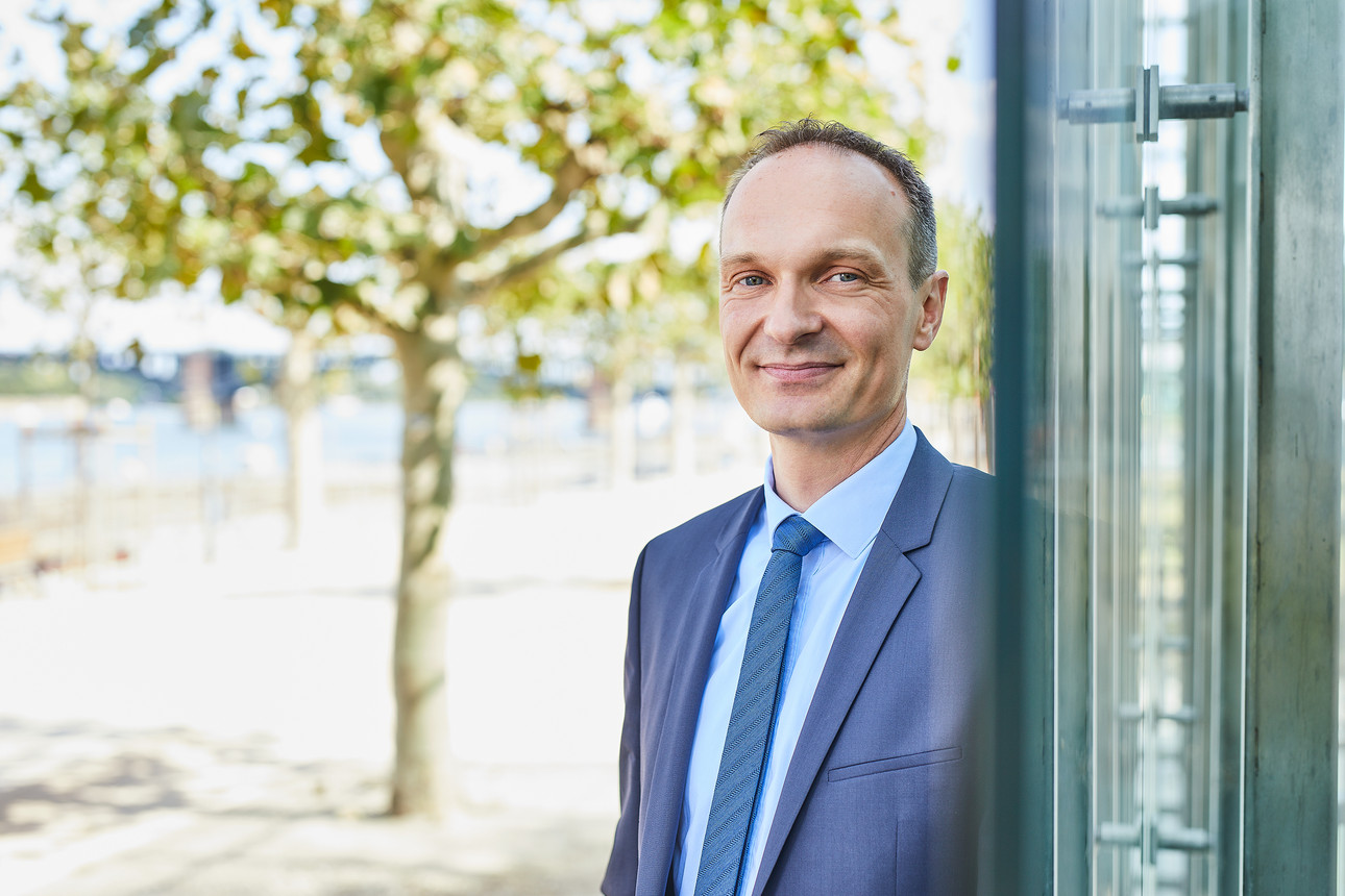 Ein Mann lehnt in Anzug und Krawatte an einer Glasfront. Er lächelt in die Kamera.