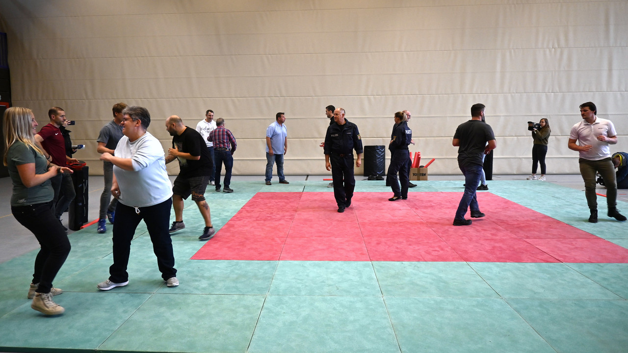 Eine Gruppe von Menschen trainiert in einer Turnhalle. Auf dem Boden sind Matten ausgelegt. 