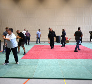 Eine Gruppe von Menschen trainiert in einer Turnhalle. Auf dem Boden sind Matten ausgelegt. 