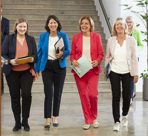 Vier Frauen schreiten durch eine doppelflügelige Tür. Sie haben Mappen in der Hand und schauen fröhlich in die Kamera.