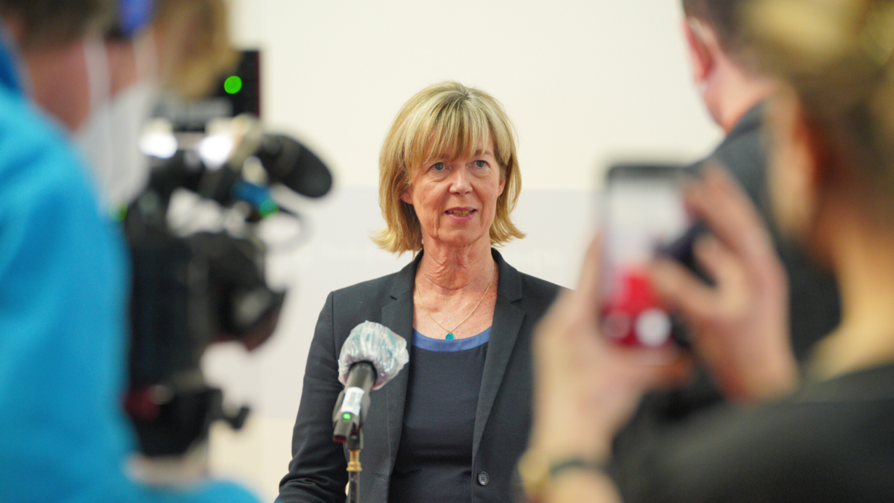 Ministerin Doris Ahnen steht vor einem Mikrofon. Sie gibt ein Interview. Mit dem Rücken zur Kamera sind drei weitere Personen unscharf zu erkennen. 