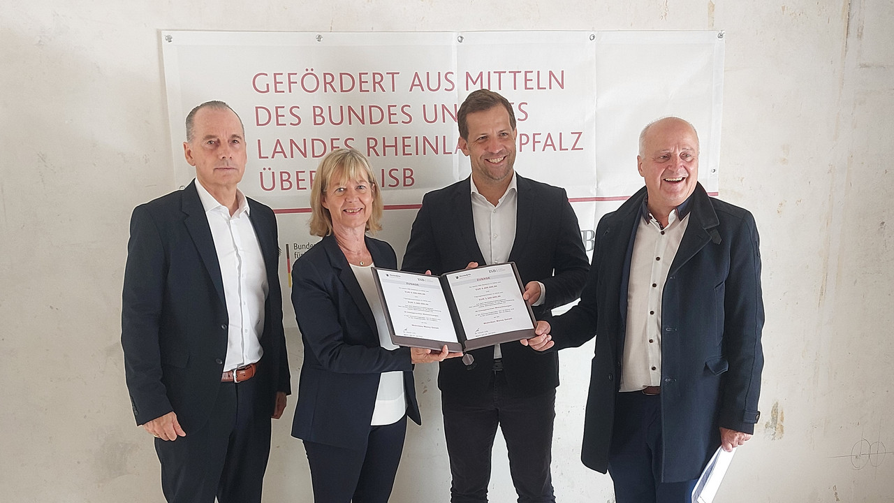 Eine Gruppe von vier Personen steht vor einem Banner. Sie halten einen Förderbescheid in der Hand.