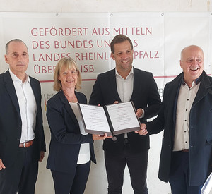 Eine Gruppe von vier Personen steht vor einem Banner. Sie halten einen Förderbescheid in der Hand.