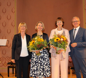 Eine Gruppe von vier Personen lacht in die Kamera. Die zwei Frauen in der Mitte halten einen Blumenstrauß in der Hand.