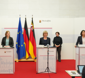 Drei Frauen stehen an Redepulten. Sie lächeln in die Kamera. Im Hintergrund sind zwei Gebärdendolmetschende platziert. 