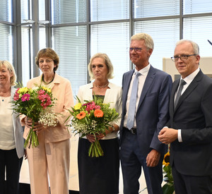 Eine Gruppe von fünf Personen steht nebeneinander. Sie lächeln. Zwei Frauen haben Blumensträuße in der Hand. 