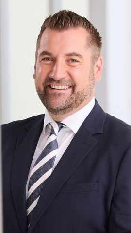 Portraitaufnahme von einem Mann im dunkelblauem Anzug mit weißem Hemd und blau-weiß gestreifter Krawatte
