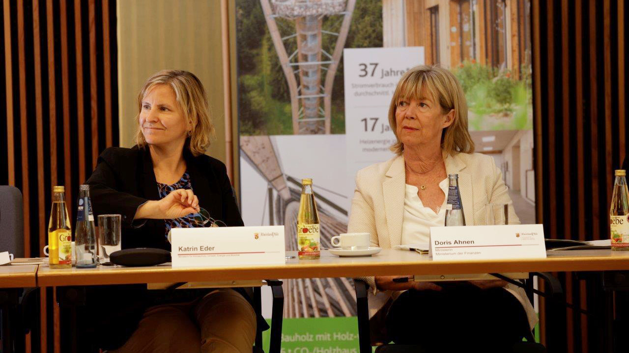 Zwei Frauen sitzen an Tischen. Vor ihnen stehen Namensschilder.